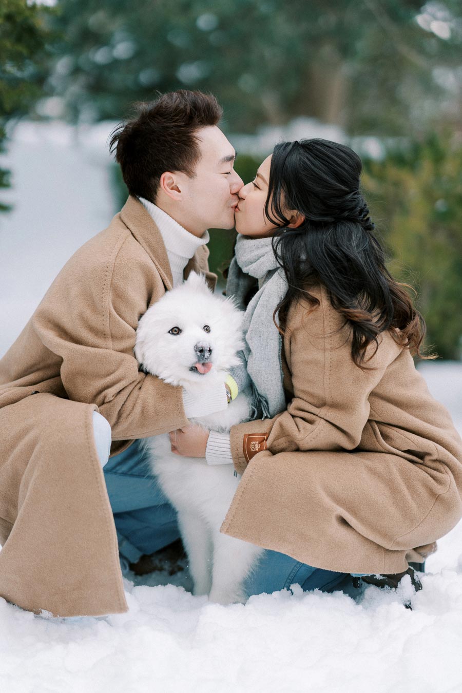 Couple kissing while holding a white dog in snowy landscape, both wearing matching beige coats, showcasing love and winter fashion.