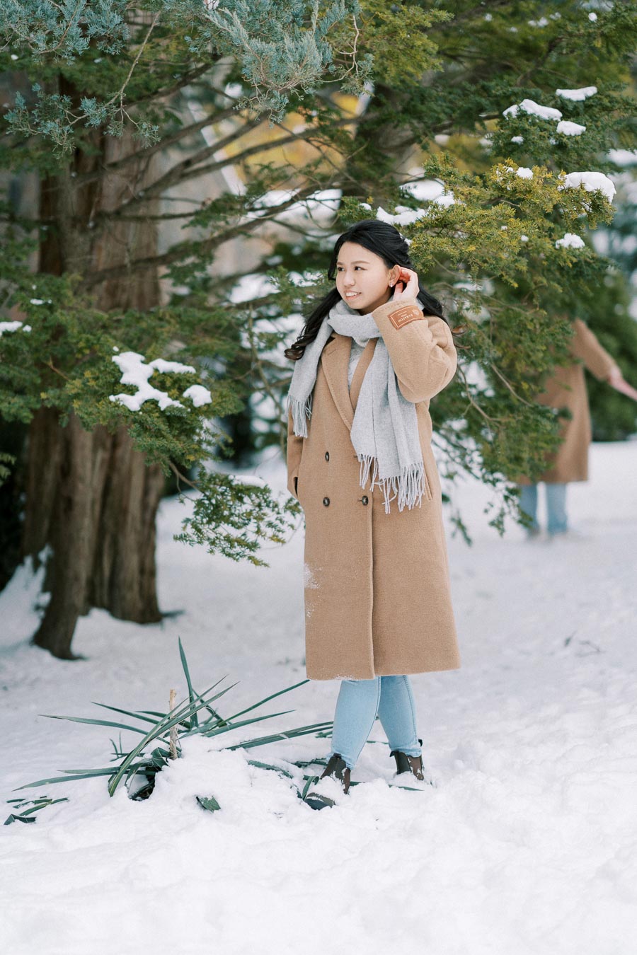 A person wearing a long camel coat and gray scarf standing in a snowy forest, surrounded by snow-covered evergreen trees.