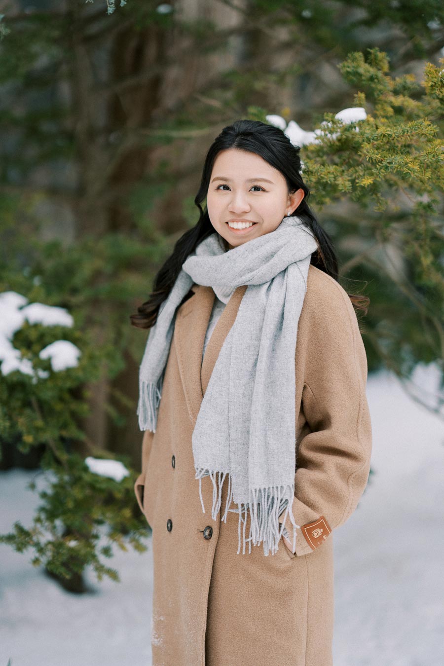 A person in a warm brown coat and gray scarf standing in a snowy landscape with evergreen trees.