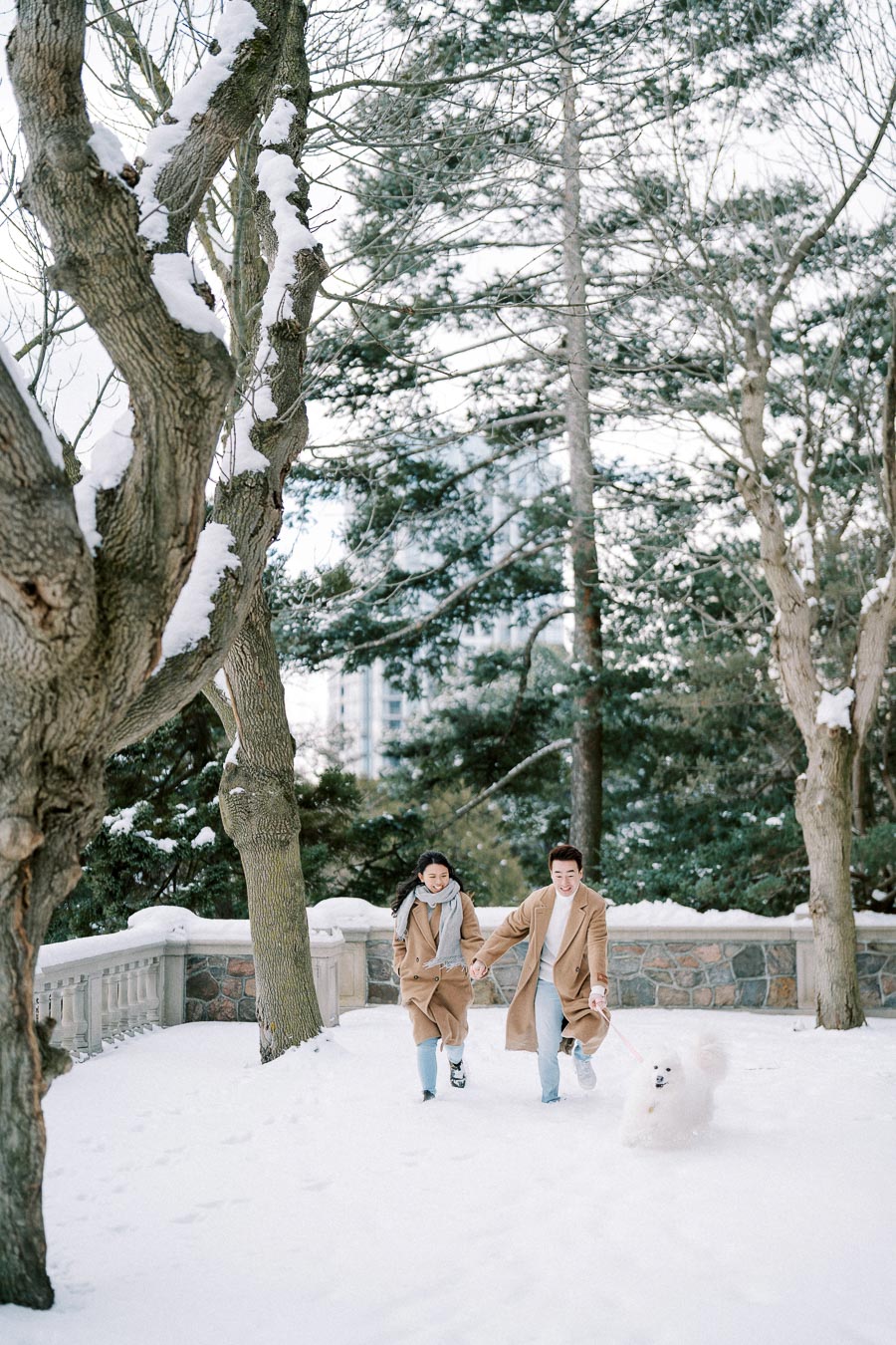 A happy couple in matching brown coats walks their fluffy white dog through a snow-covered park, surrounded by snow-dusted trees, under a clear winter sky.
