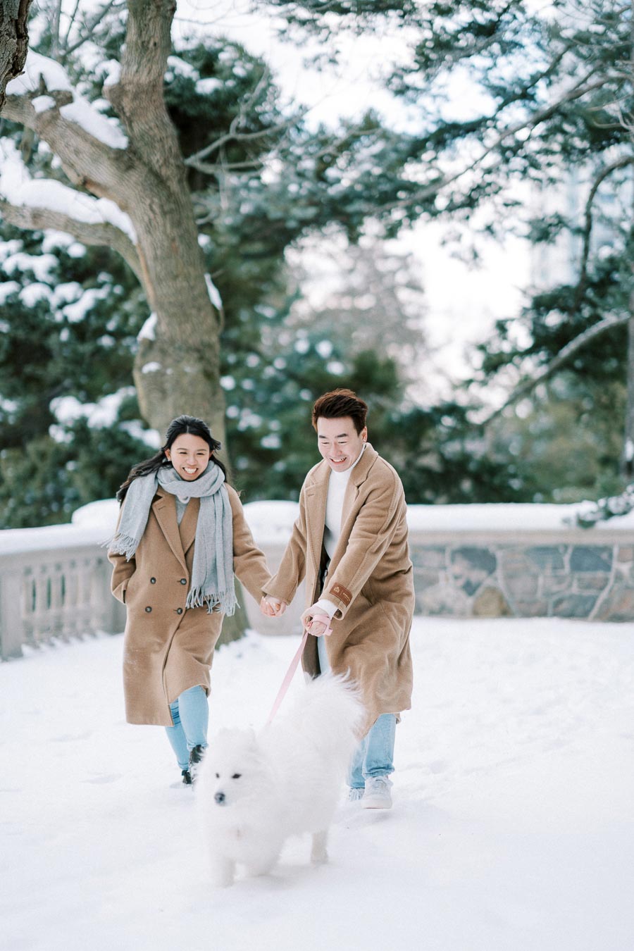 A happy couple walks their fluffy white dog through a snowy park, both wearing cozy tan coats and scarves, with snow-covered trees in the background.