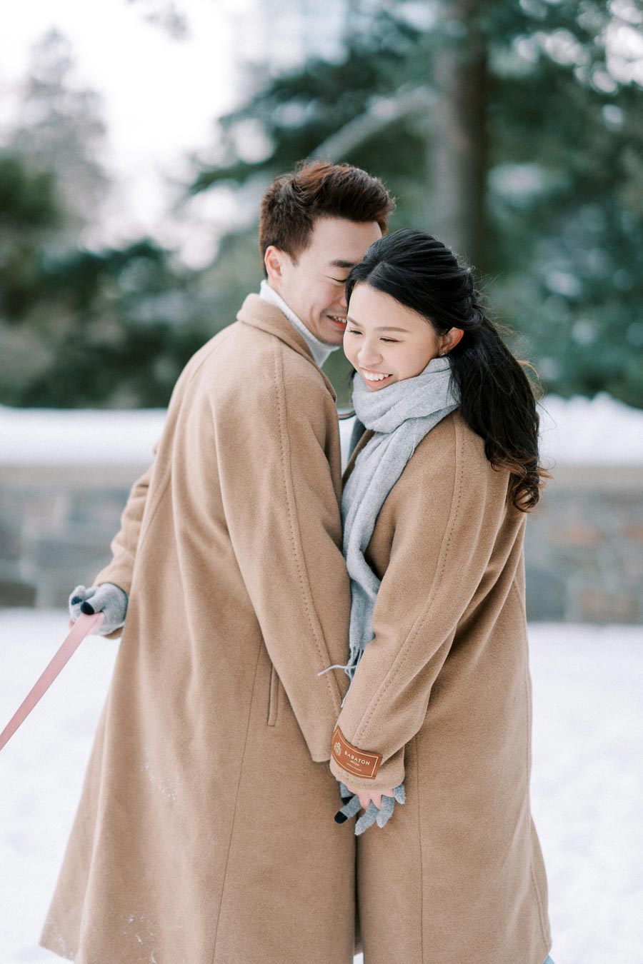 A couple smiling and embracing in a snowy park while wearing matching tan coats and gray scarves, surrounded by blurred trees.
