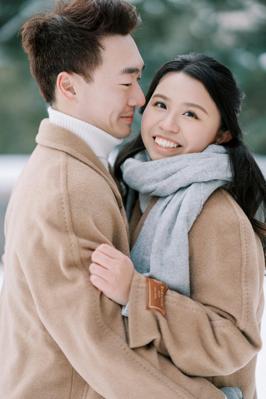 A couple happily embracing in a winter setting, wearing beige coats and a gray scarf, with a snowy background.