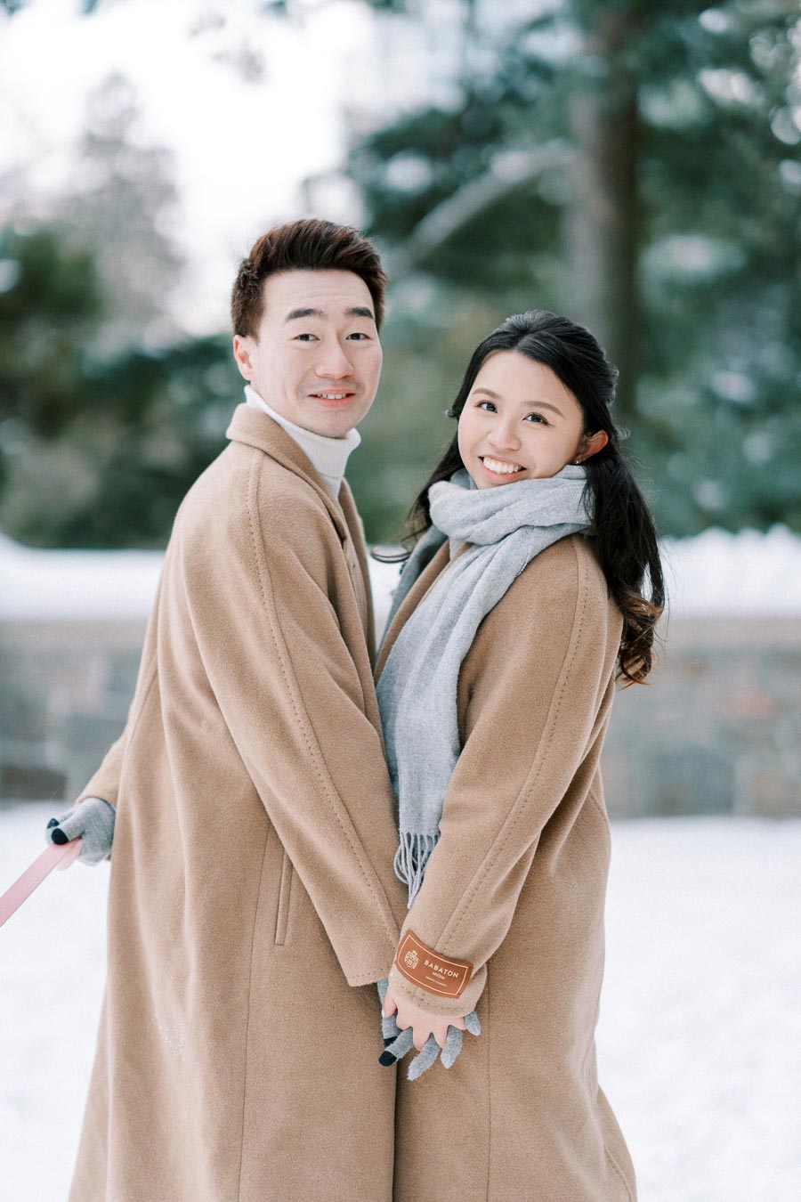Couple smiling while holding hands in winter coats in a snowy park.