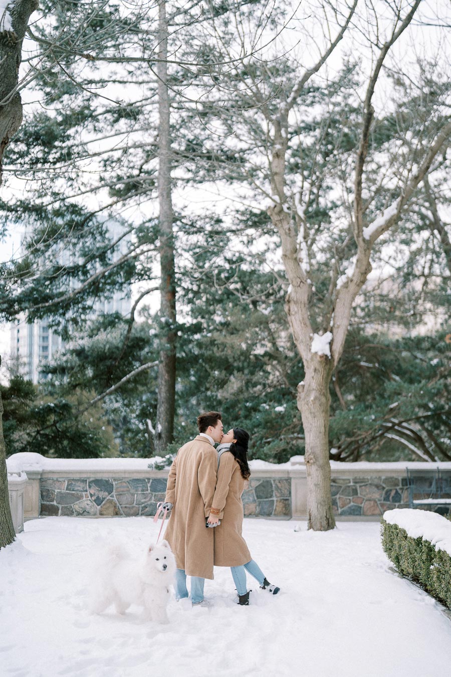 A couple in matching tan coats shares a kiss in a snowy park, accompanied by a fluffy white dog on a leash.