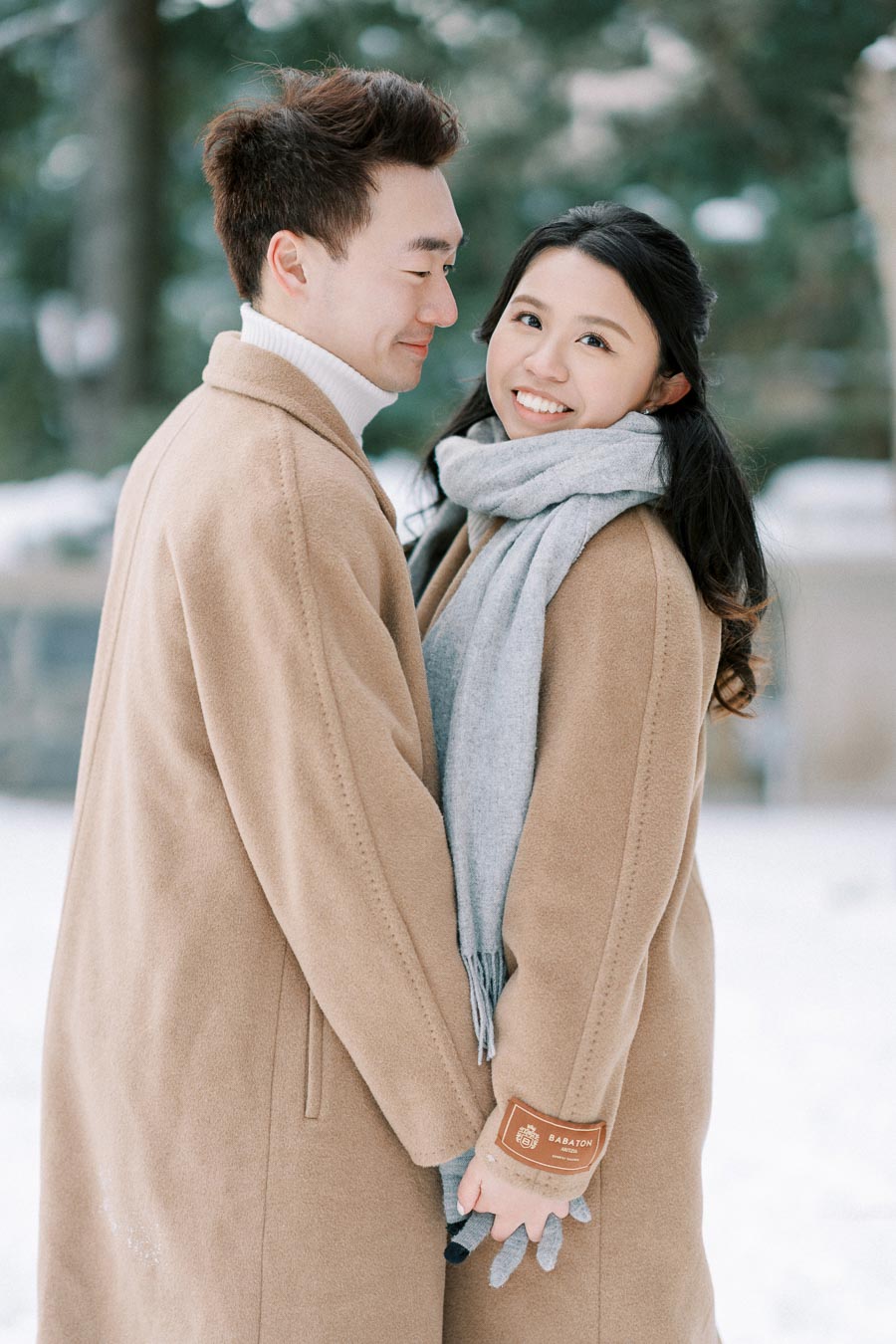 A couple wearing beige coats and a gray scarf smiles while standing in a snowy winter landscape, holding hands and gazing at each other affectionately.