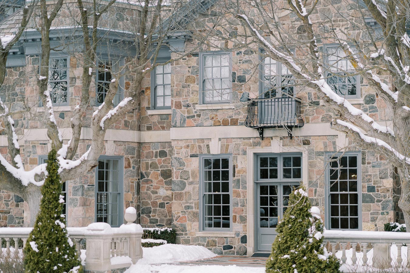 Stone house surrounded by snow-covered trees and shrubs in a winter setting.