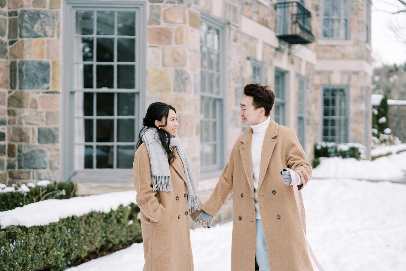 Couple wearing beige coats, holding hands and smiling at each other, walking outside a stone building in a snowy winter setting.