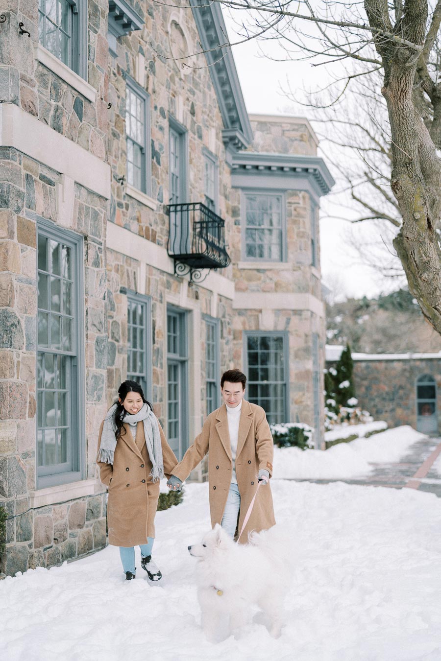 A couple holding hands, dressed in matching tan coats, strolls outside a stone building on a snowy day, accompanied by a fluffy white dog.