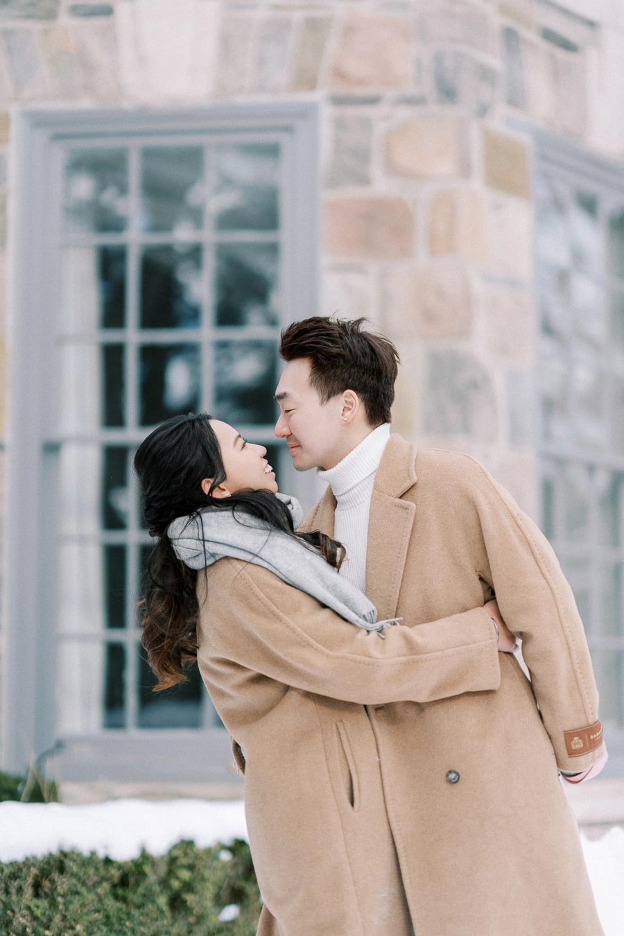 Couple embracing in winter coats outside a stone building with large windows, set against a snowy backdrop.