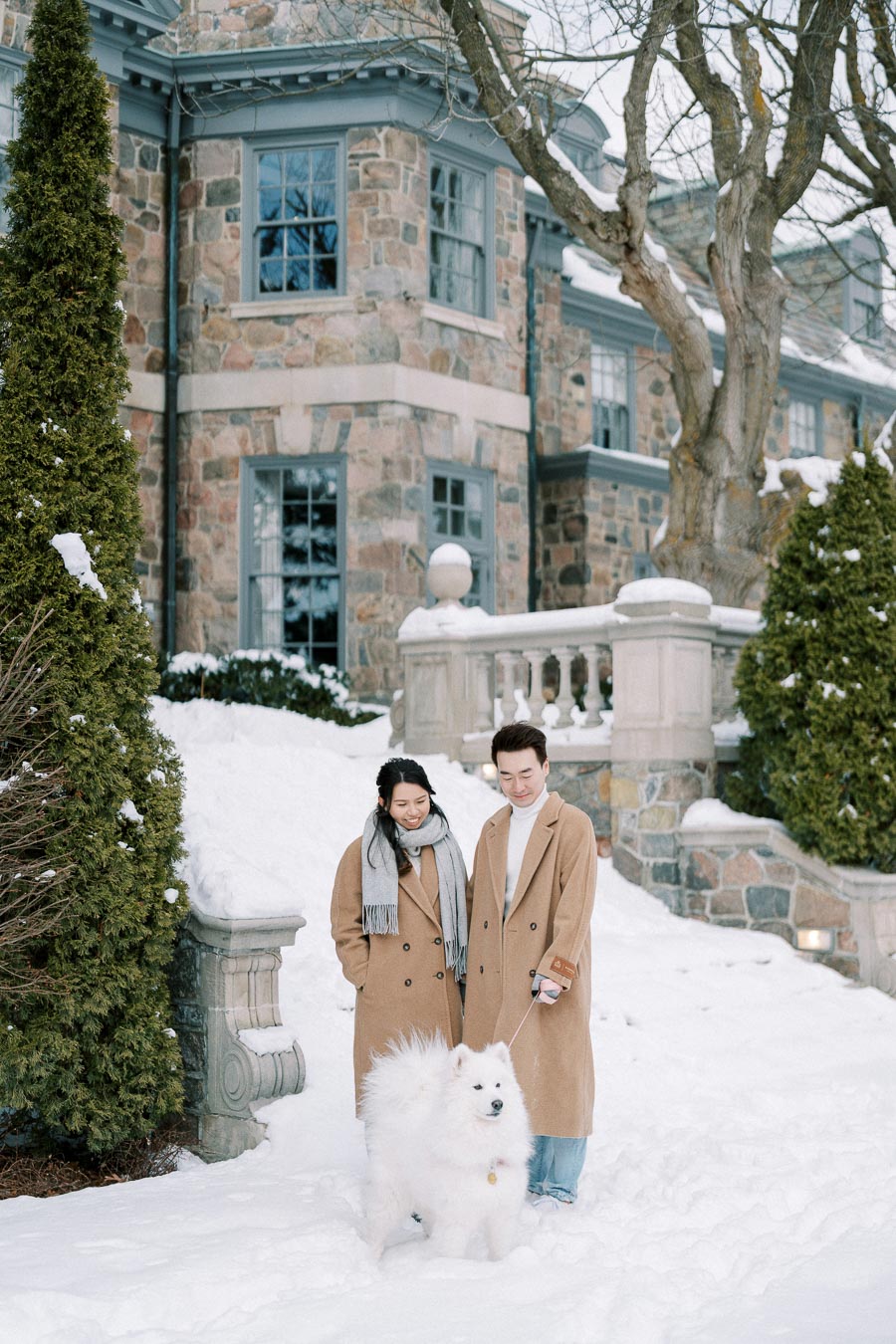 A couple dressed in winter coats standing in front of a stone house on a snowy day, smiling while holding the leash of their fluffy white Samoyed dog.
