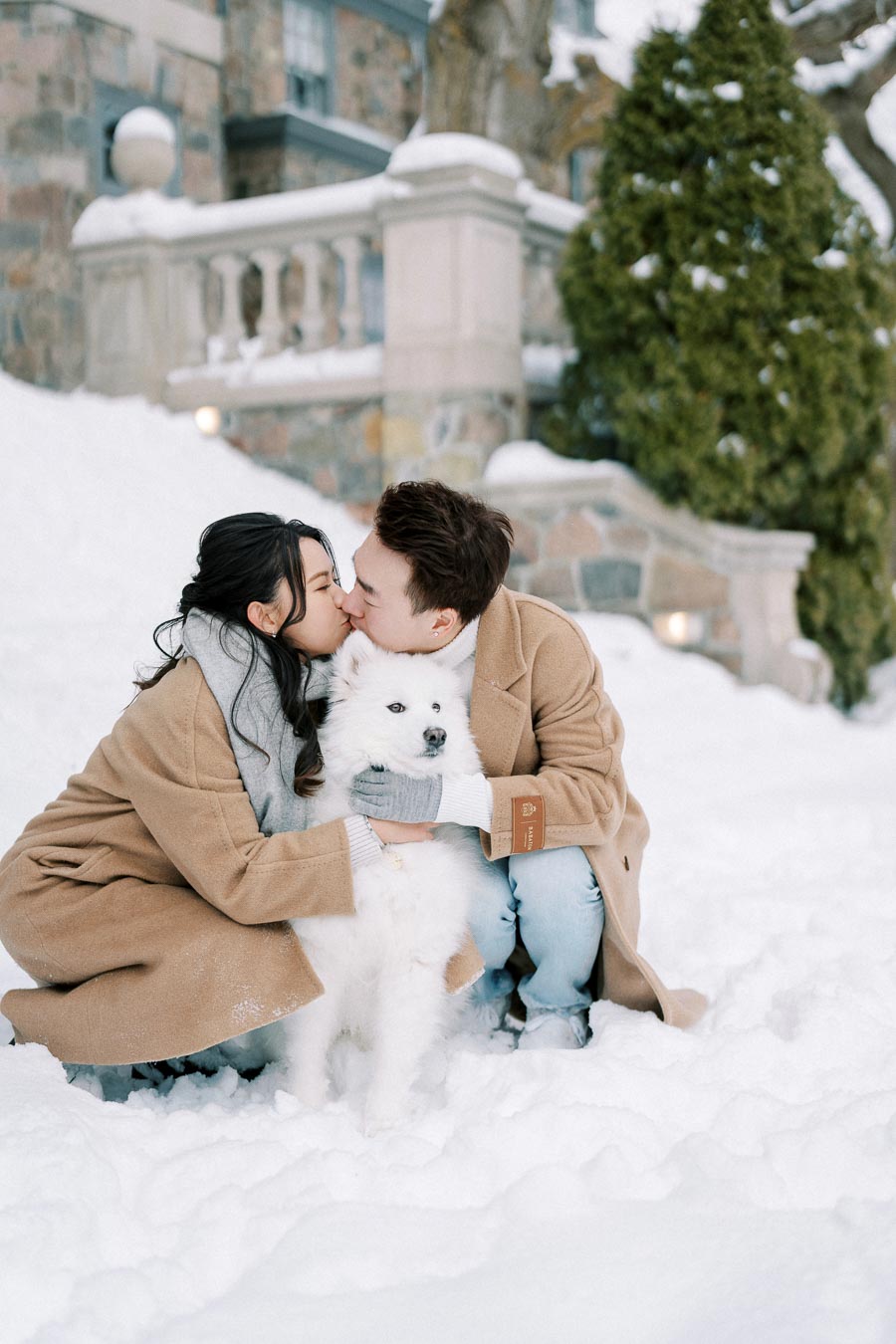 A couple in beige coats kissing while cuddling their fluffy white dog in a snowy winter setting, with a stone building and evergreen trees in the background.