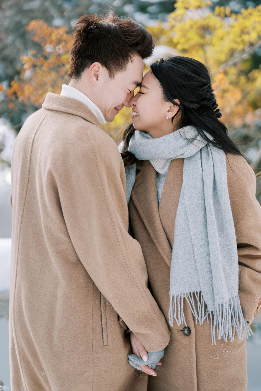 A couple in warm coats and scarves shares a loving moment by touching foreheads in a snowy autumn landscape.