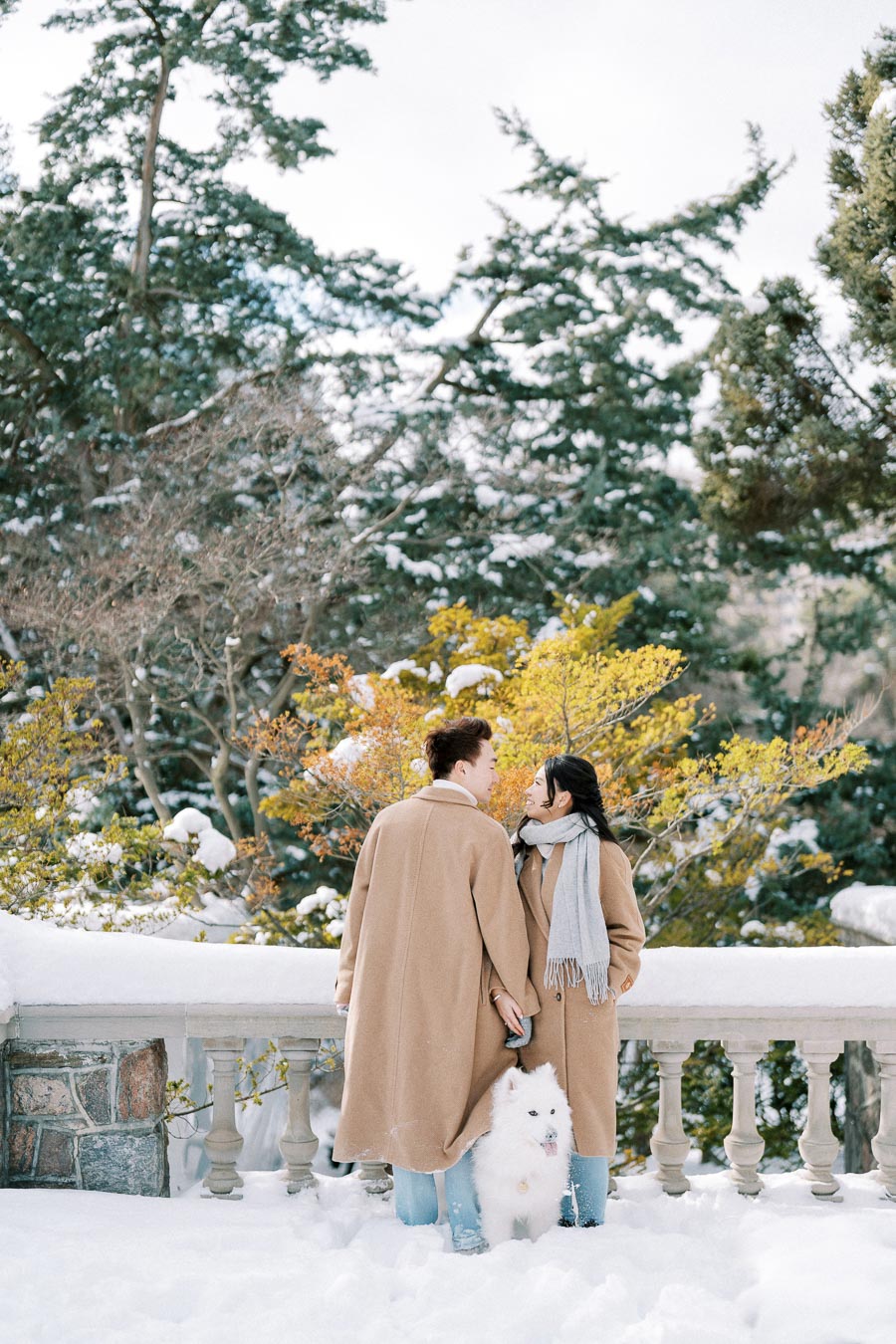 Couple in beige coats with a white dog standing on a snowy path, surrounded by winter foliage and trees.