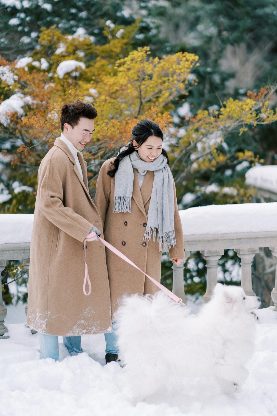 A couple in matching beige coats and gray scarves walking a fluffy white dog in a snowy park, surrounded by snow-covered trees and colorful foliage.