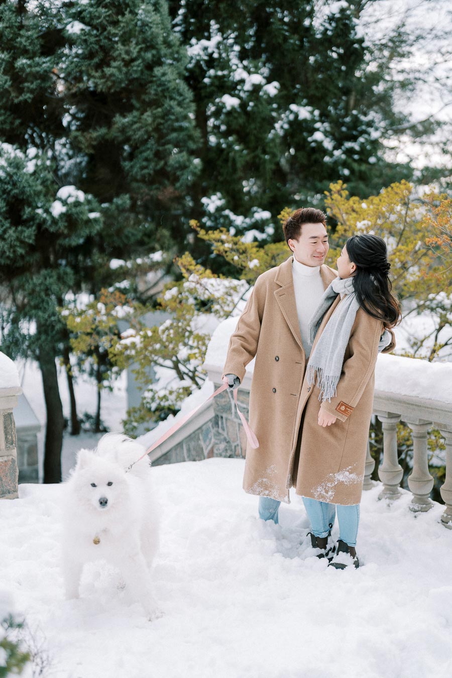 Couple in winter coats smiling at each other while walking their white fluffy dog on a snow-covered path, surrounded by evergreen trees.