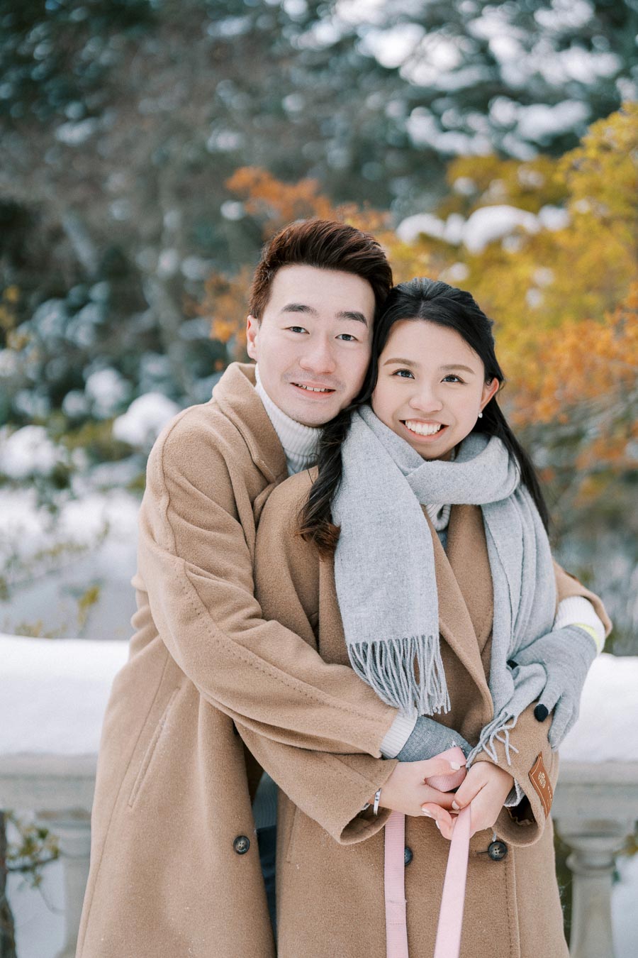 A happy couple embracing in a snowy winter landscape, both wearing warm brown coats and grey scarves, with a backdrop of snow-dusted trees and orange foliage.