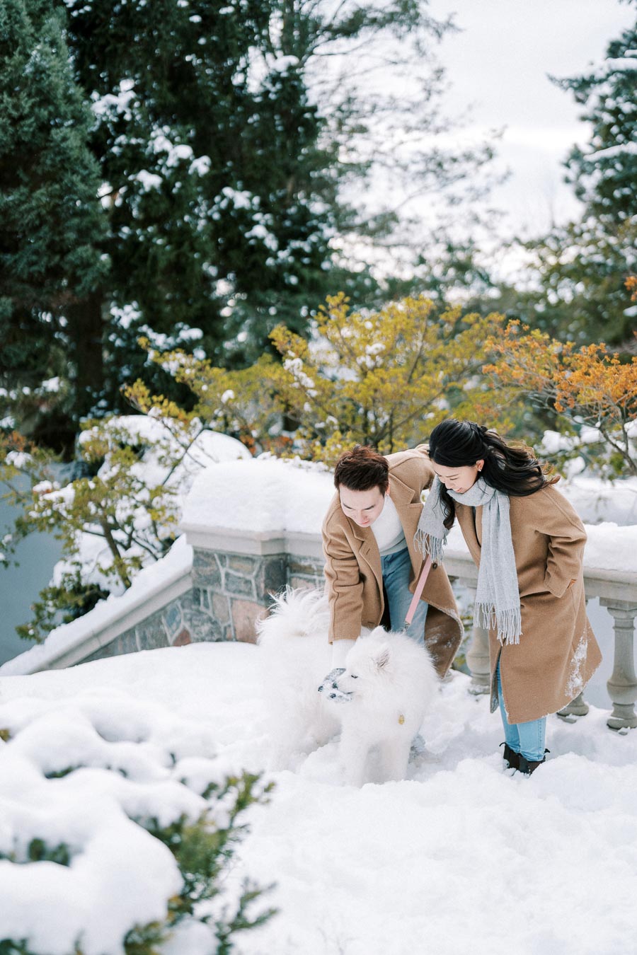 A couple in matching brown coats kneels in a snowy garden, petting a fluffy white dog. Surrounding the scene are snow-covered trees and shrubs, creating a peaceful winter atmosphere.