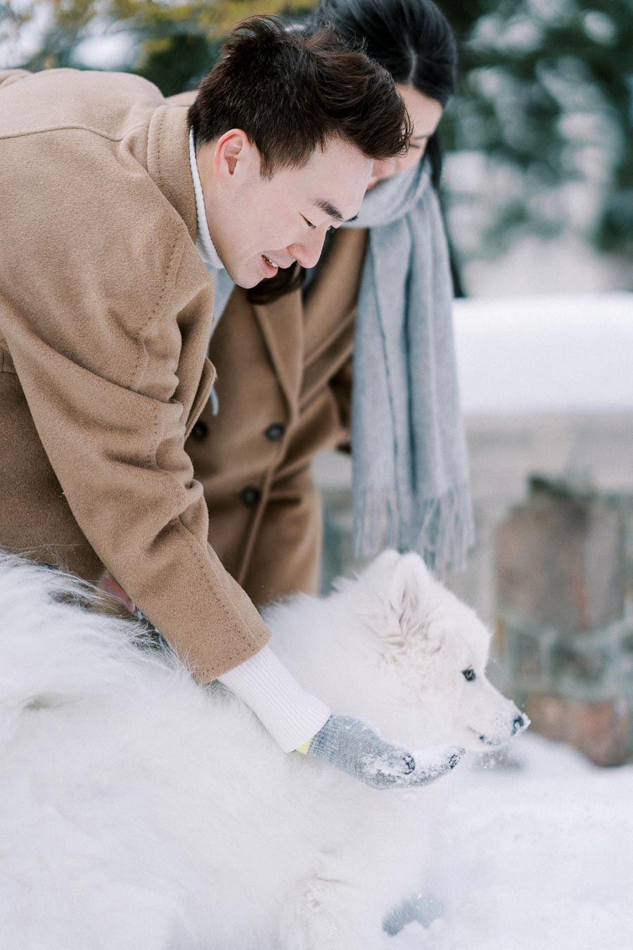 Two people in winter coats petting a fluffy white dog in the snow.