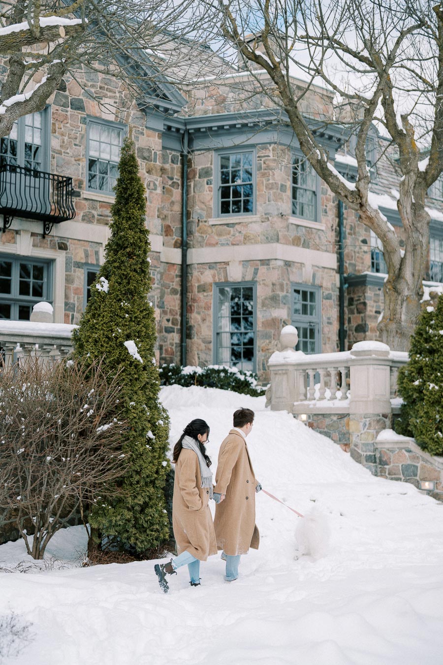 Couple walking their dog in the snow outside a large stone house, wearing matching tan coats and scarves on a winter day.