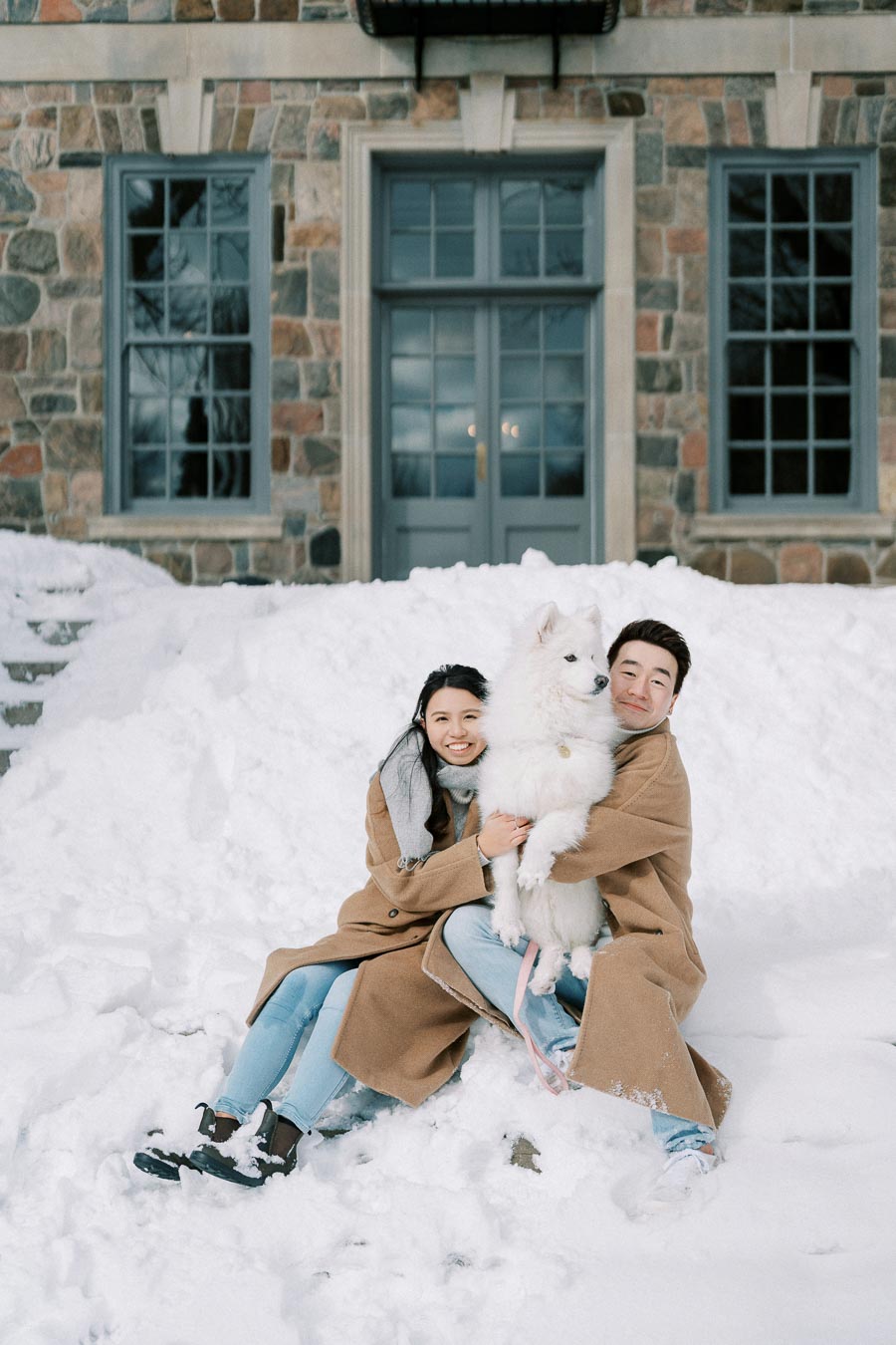 Couple enjoying a snowy day with their white dog outside a stone building.