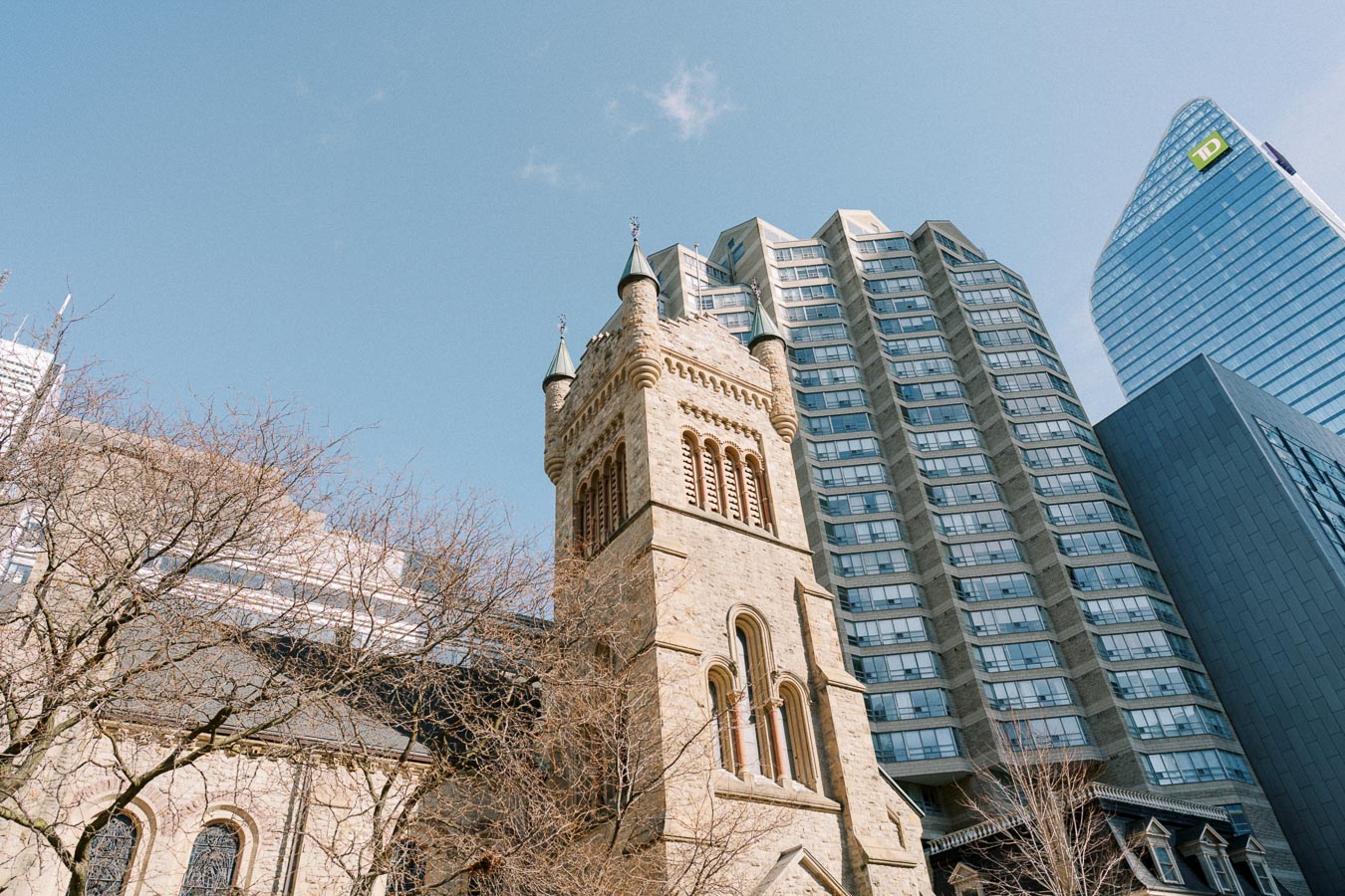 A historic stone church tower stands in contrast to modern skyscrapers in an urban setting, under a clear blue sky.