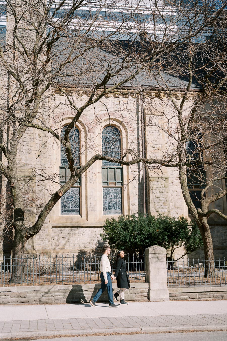 A couple walks hand in hand past a historic stone building with arched windows and intricate stained glass, framed by bare trees.