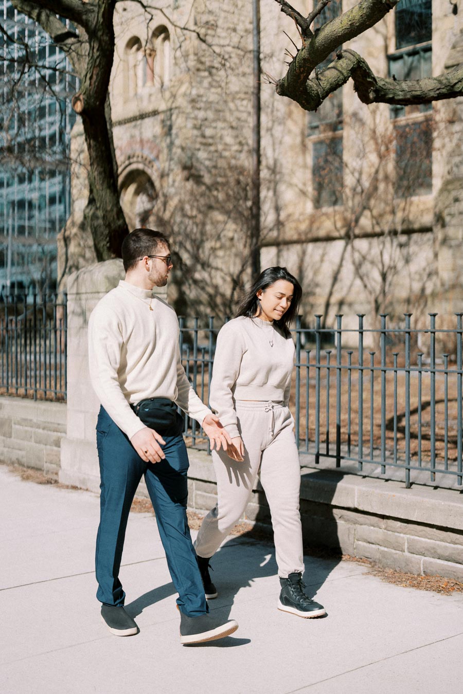 A man and woman walk together on a sunny day, passing by a historic stone building and iron fence.