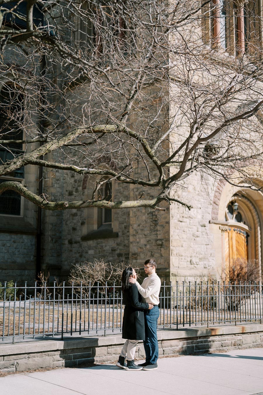 A couple shares a tender moment in front of a historic stone building, with bare tree branches framing the scene.
