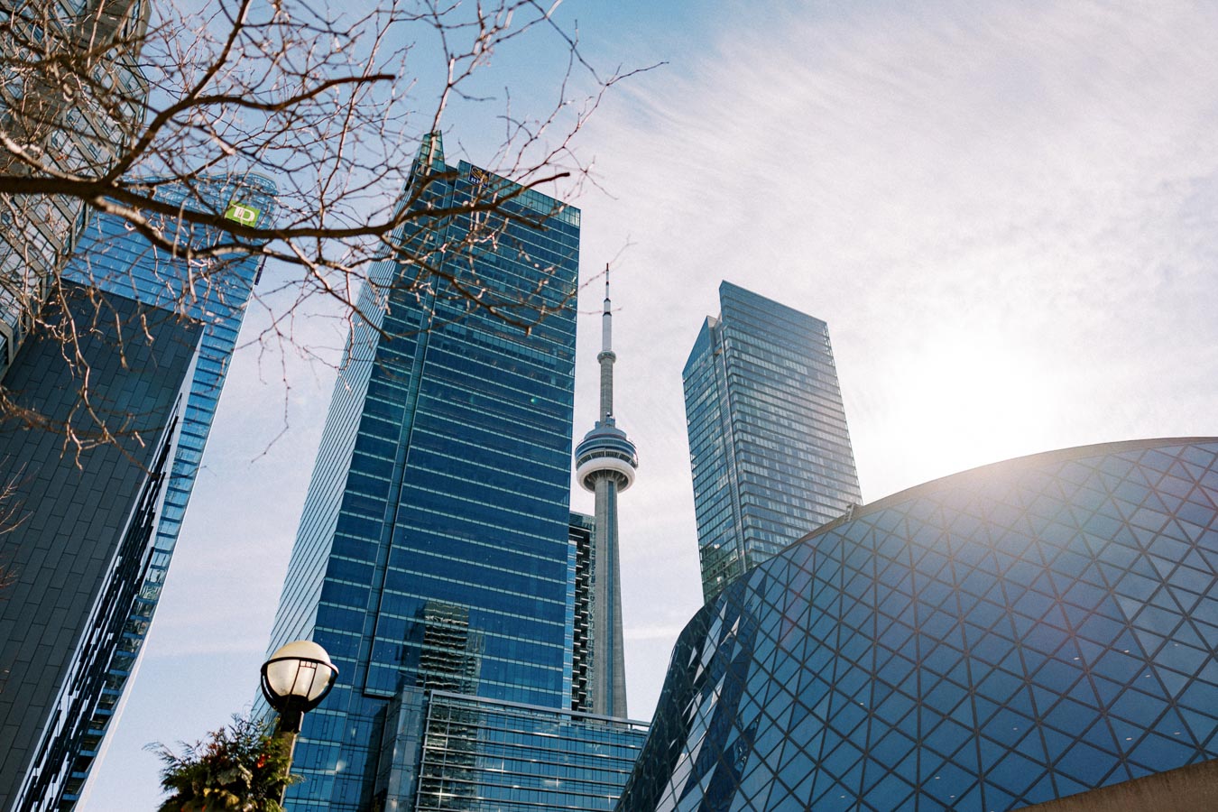 The CN Tower stands tall among modern skyscrapers in Toronto, Canada, under a clear blue sky with the sun shining brightly.