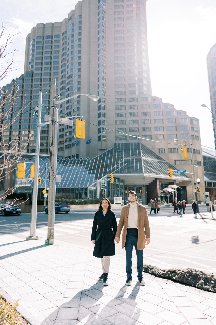 A couple walks hand in hand on a city street, with a tall modern building in the background. The scene is bright and sunny, with other pedestrians and cars visible in the urban setting.