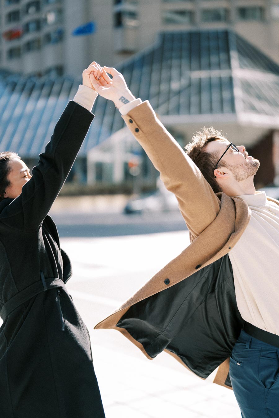 Two individuals joyfully stretch and hold hands outdoors, dressed in stylish coats, with a modern architectural structure in the background.