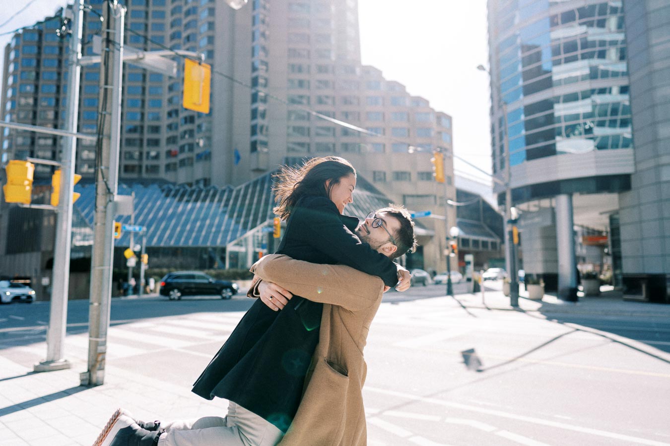 A joyful couple embraces on a city street, surrounded by modern architecture and bright sunlight.