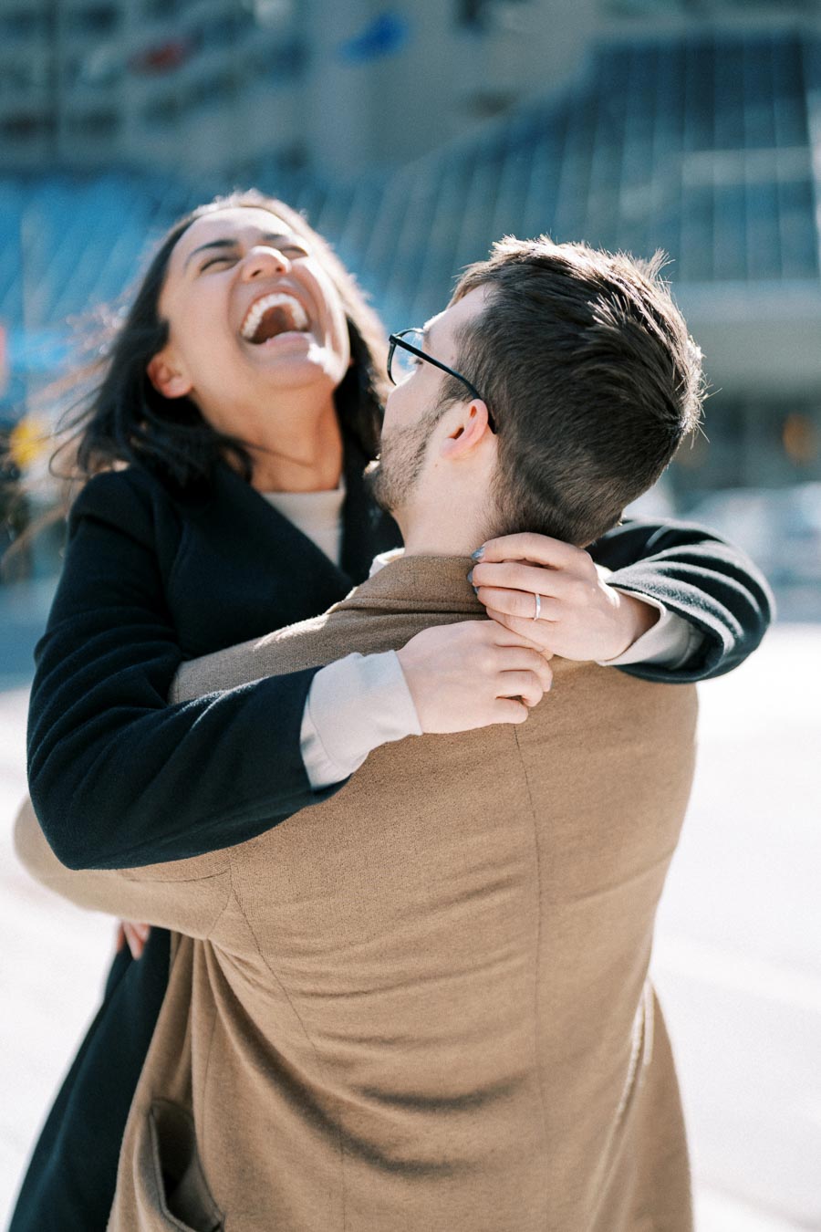 A joyful couple shares a moment of laughter while embracing outdoors, with the woman wearing a black coat and the man in a brown coat.
