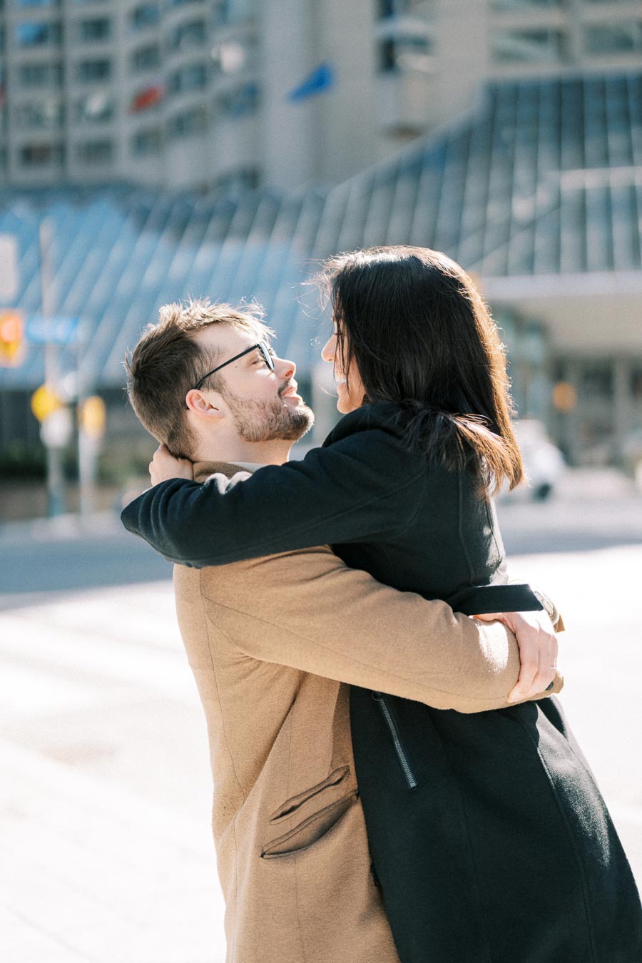 A couple shares a joyful embrace in an urban setting, with modern architecture in the background.