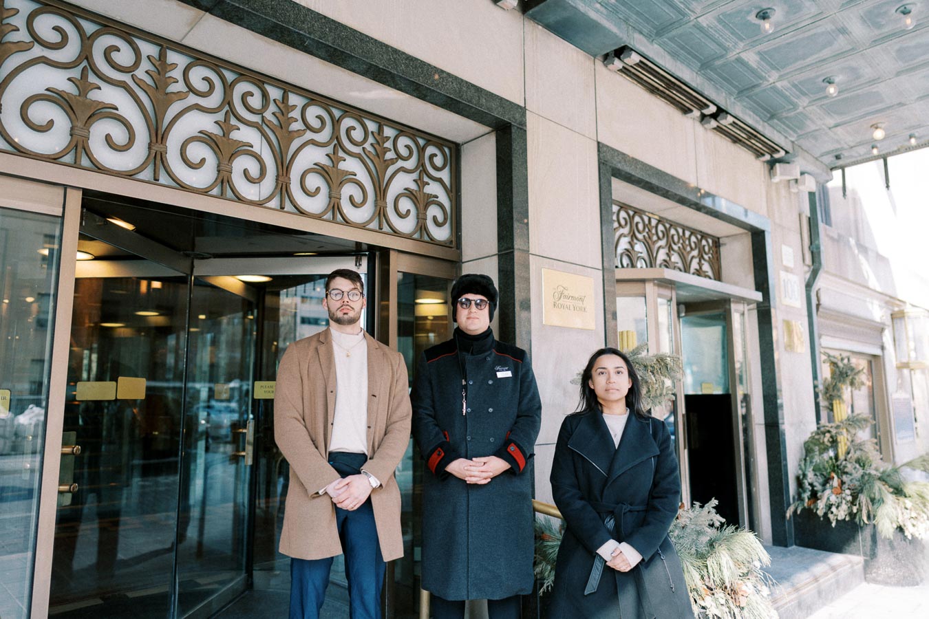 Three individuals stand in front of the Fairmont Royal York hotel entrance, showcasing its elegant design and decorative metalwork.