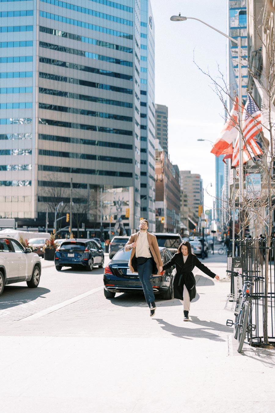 Two people joyfully jump on a city street, surrounded by tall buildings and flags, capturing a moment of happiness in an urban setting.