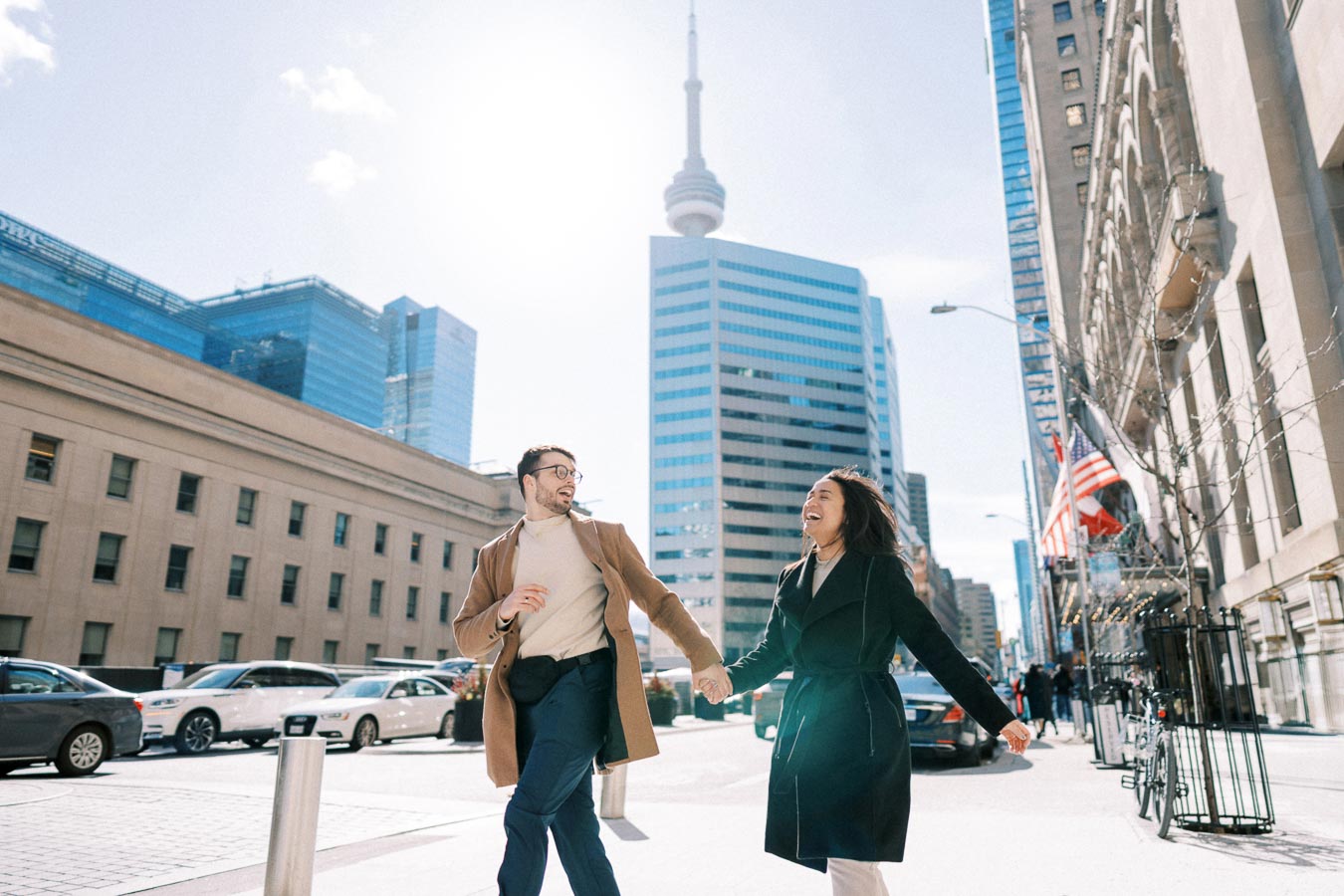 A couple joyfully walks hand in hand in a bustling city street with the iconic CN Tower visible in the background, symbolizing a lively urban atmosphere.