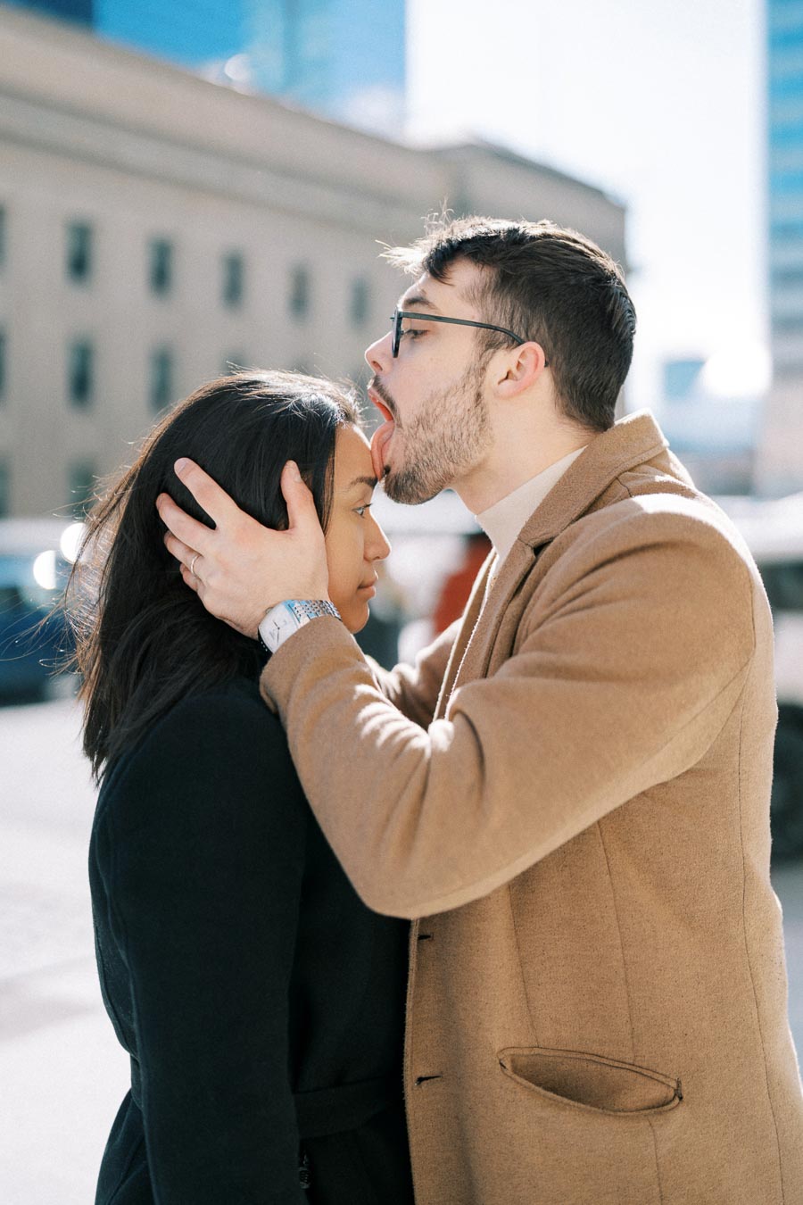A playful moment captured between a man and a woman on a city street, with the man in a tan coat playfully licking the woman's forehead.