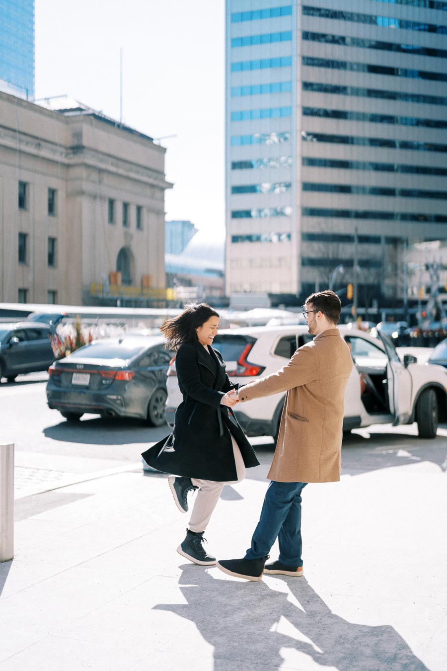 A couple joyfully dances on a city sidewalk, surrounded by urban architecture and parked cars.