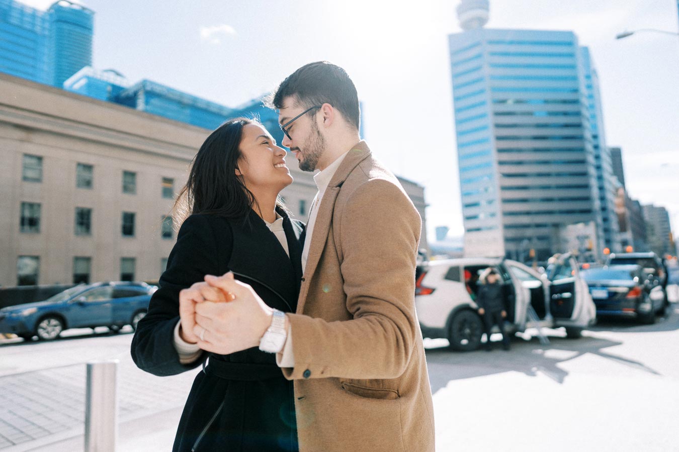 A couple shares a joyful moment on a sunny city street, surrounded by modern buildings and cars.
