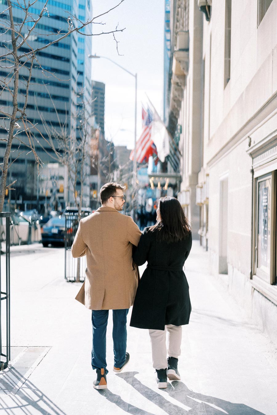 A couple strolls down a city street on a sunny day, surrounded by tall buildings and flags.