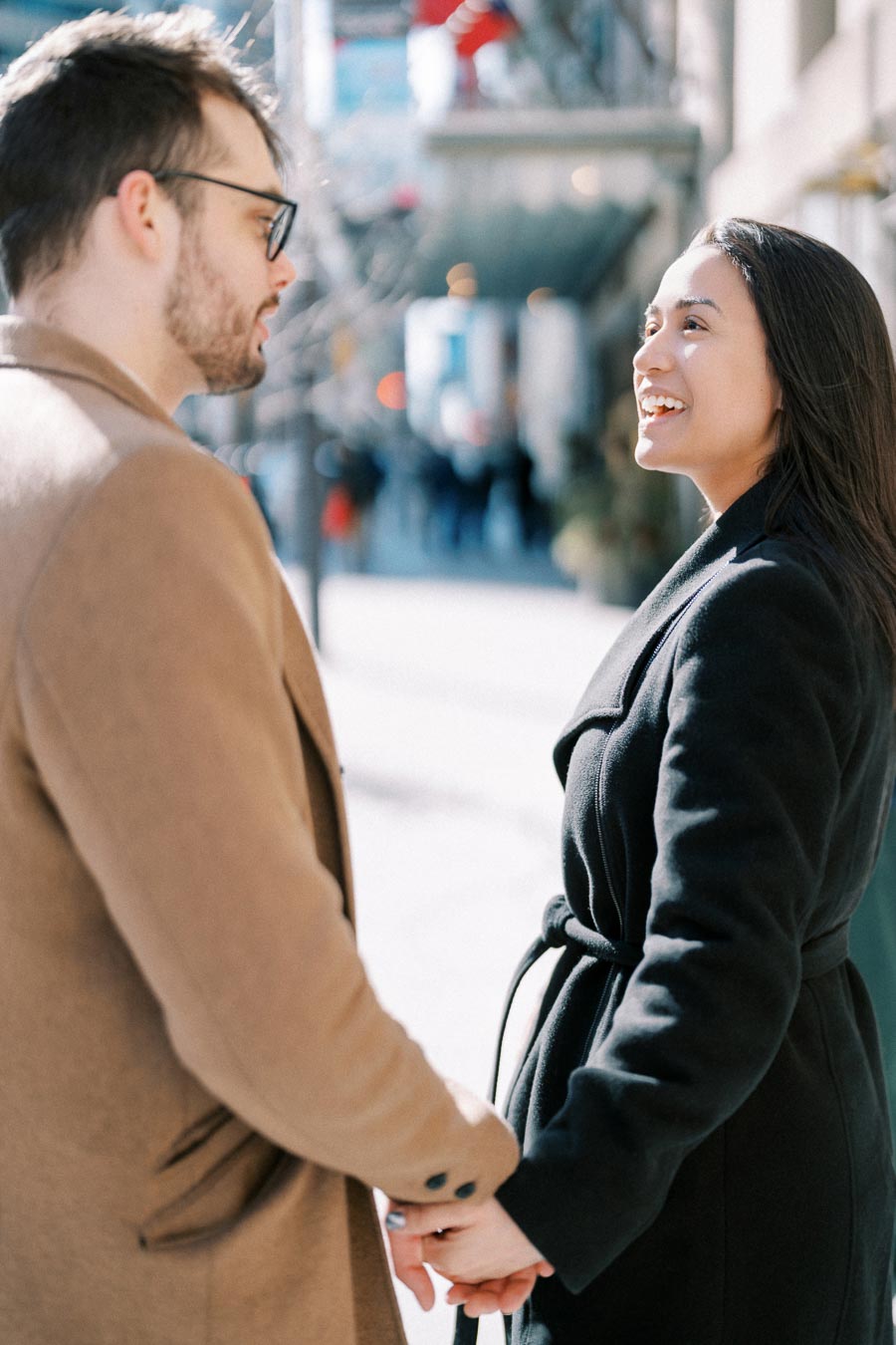 A couple shares a joyful moment on a sunny day, holding hands and smiling at each other on a city street.
