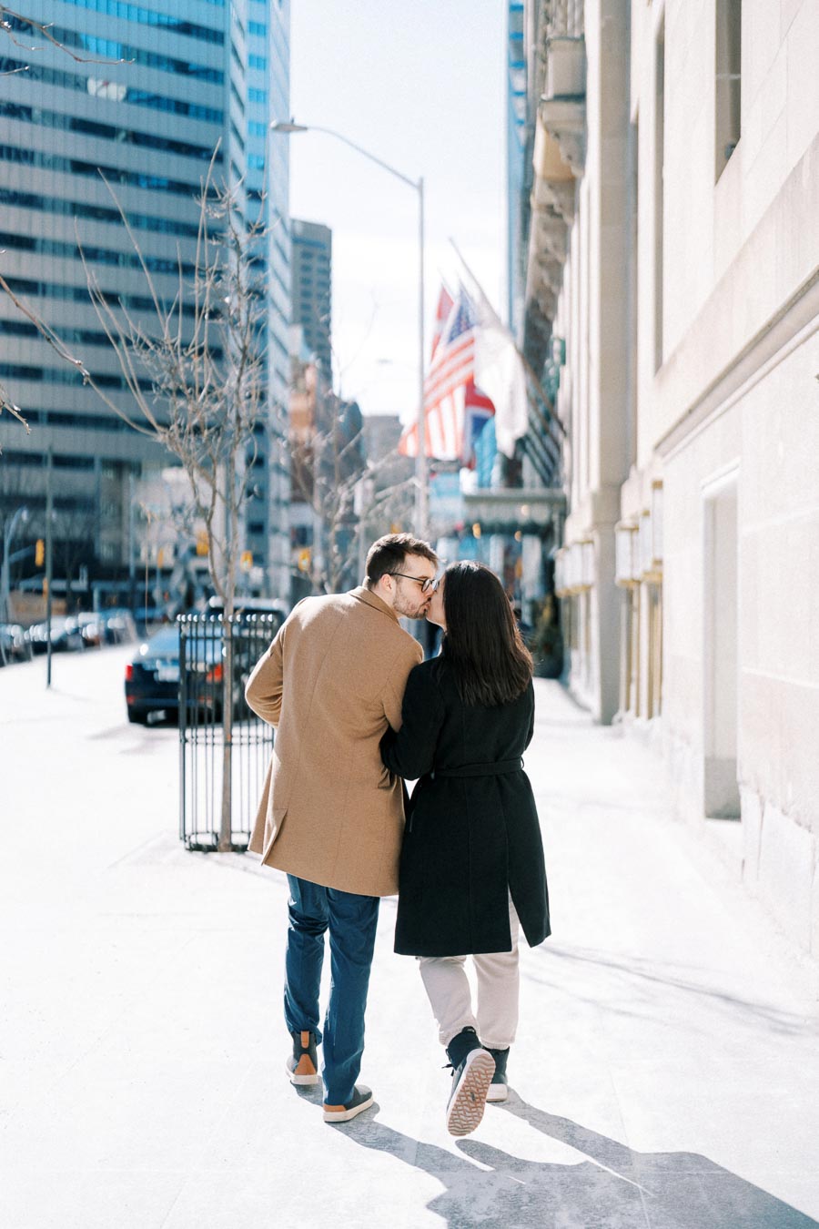 A couple shares a kiss while walking down a city sidewalk, surrounded by tall buildings and American flags.