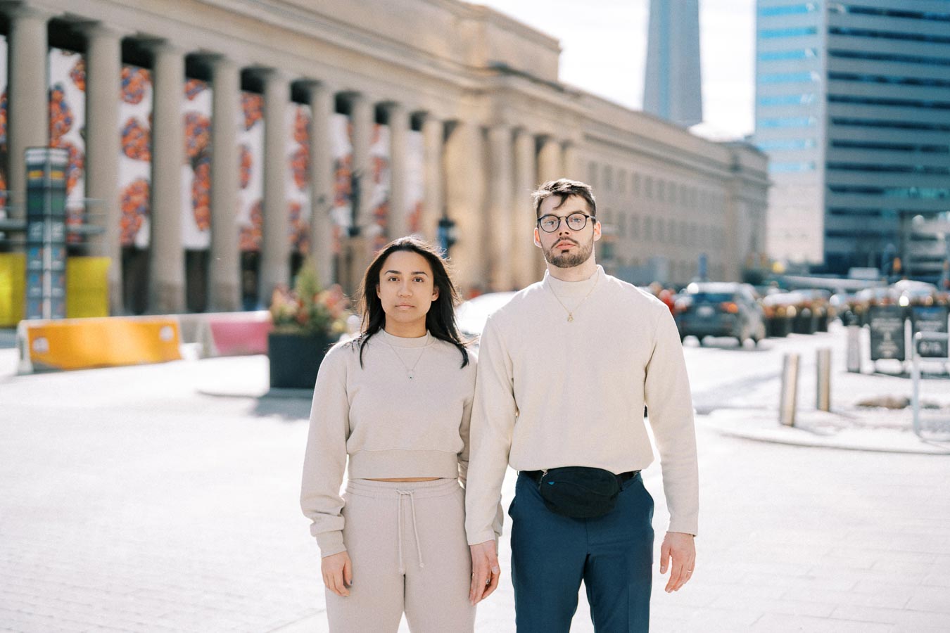 A man and woman stand together in front of a columned building in an urban area, dressed in casual attire.
