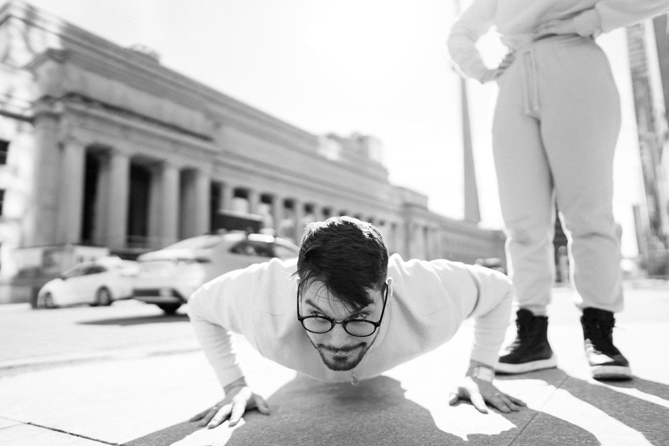A man performs push-ups on a city street, with a historic building and cars in the background.