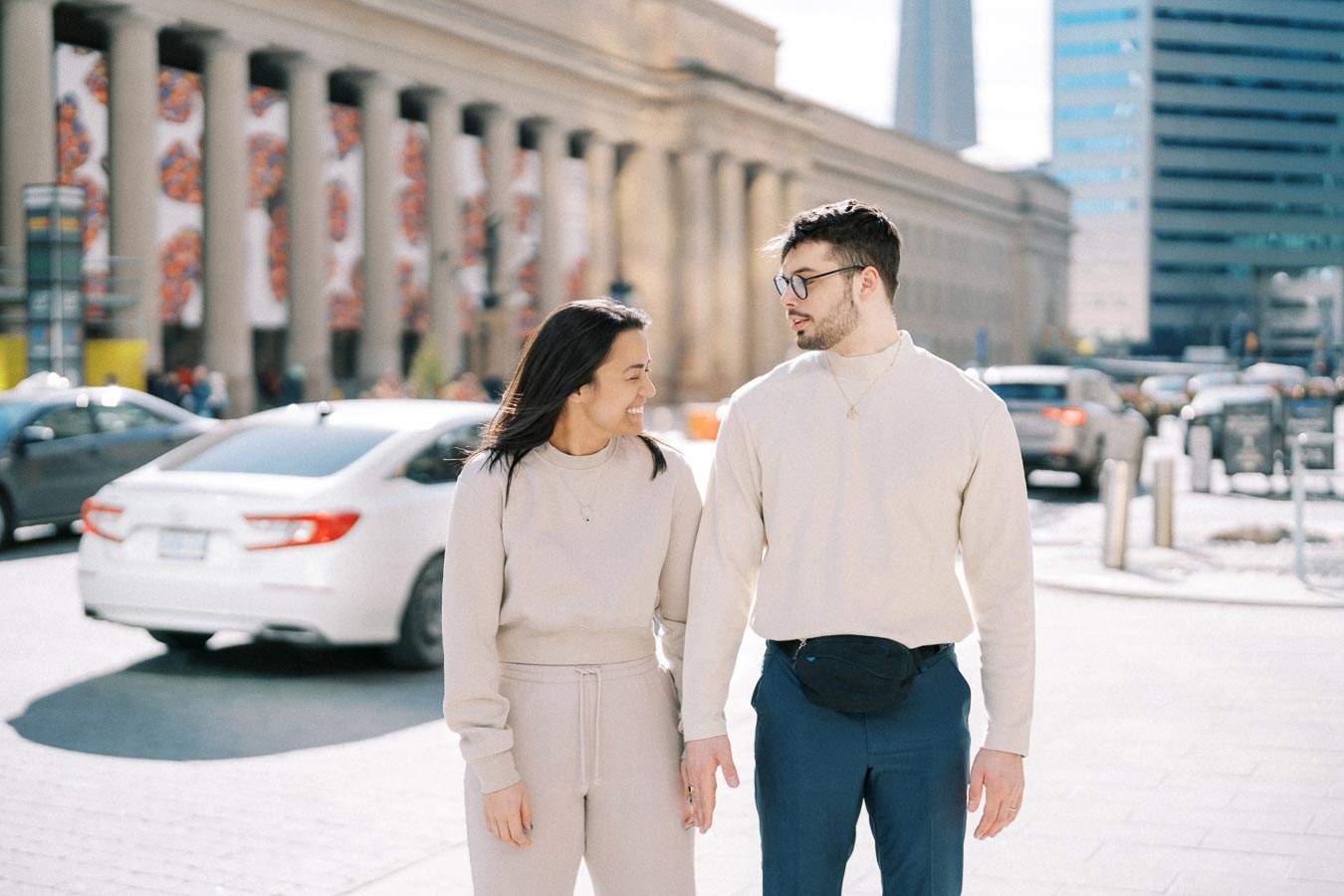 A couple walks hand in hand on a sunny day in the city, with a large building and cars in the background.