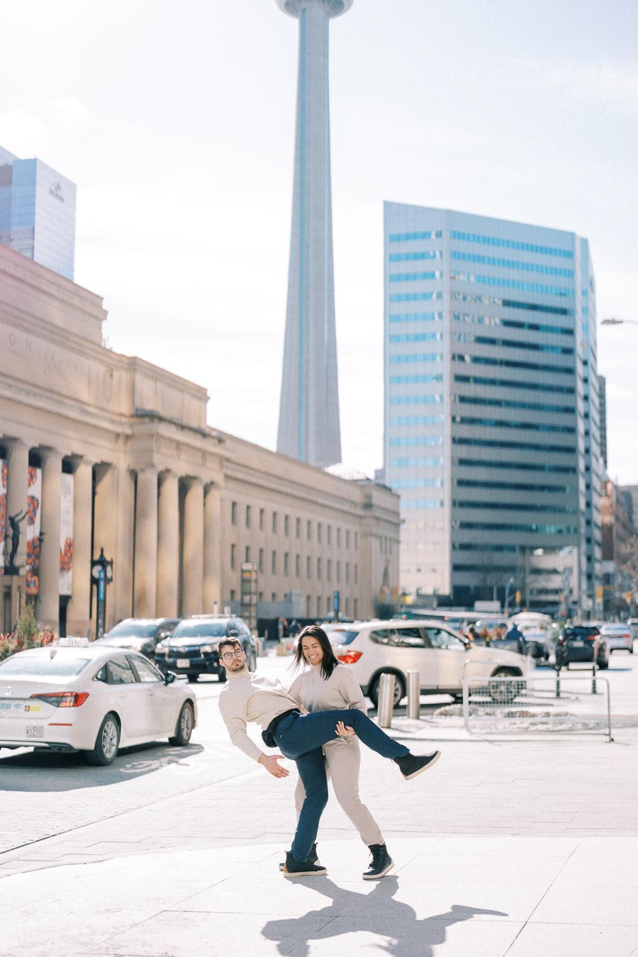 A playful couple poses in front of the iconic CN Tower in Toronto, capturing a fun moment amidst the city's bustling streets.