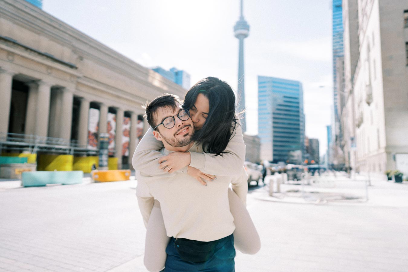 A joyful couple shares a playful moment with a piggyback ride in front of the CN Tower in Toronto.