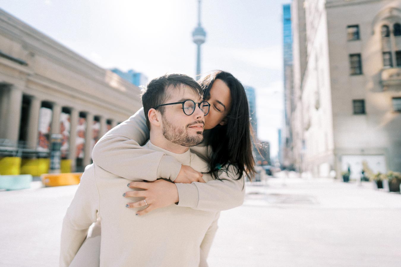 A couple shares a tender moment in a cityscape with the iconic CN Tower visible in the background, suggesting a location in Toronto.